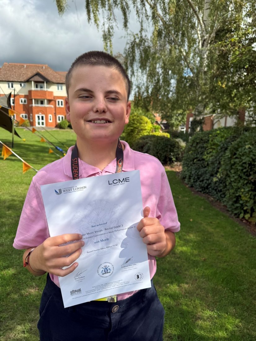 Lawson stands outdoors on a grassy area holding an LCME (London College of Music Examinations) certificate. Lawson is wearing a pink polo shirt and a student lanyard. The certificate shows a Merit in Popular Music Vocals Recital Grade 3. Trees and shrubs surround the area, with a red-brick building visible in the background. The photograph is taken in daylight with dappled shade from overhanging branches.