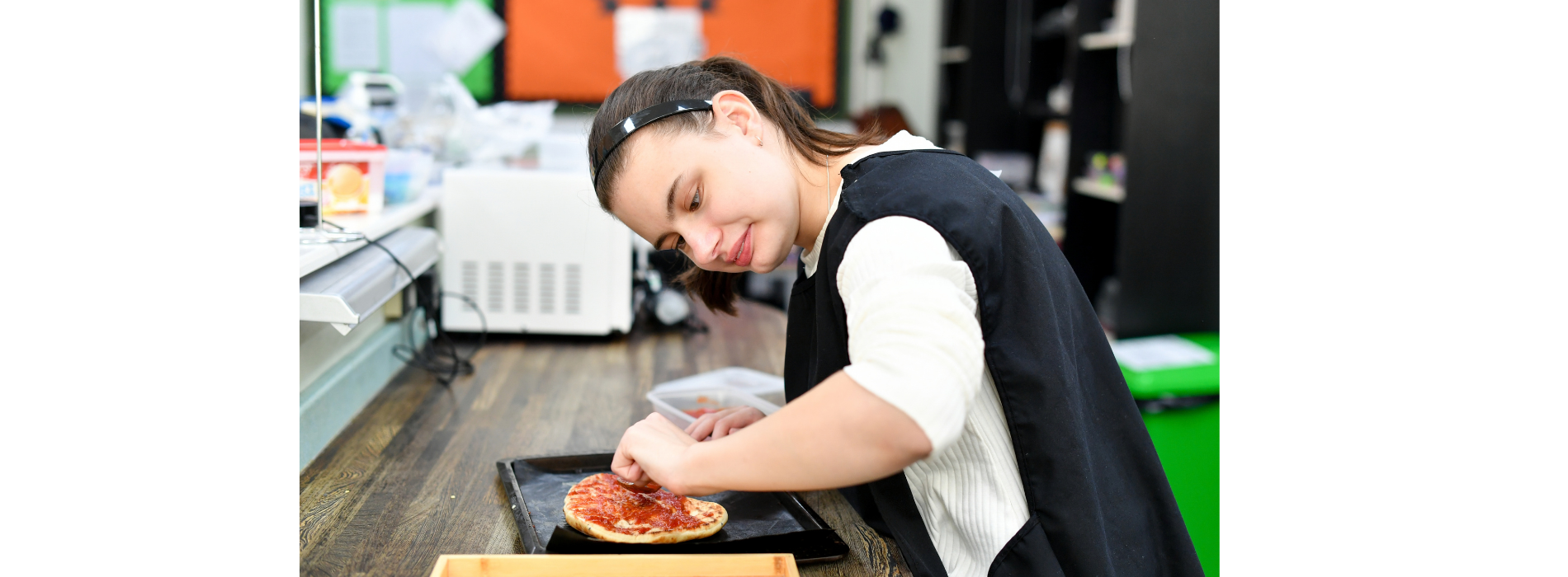 A student takes part in a practical food technology lesson, sitting at a kitchen worktop while preparing a pizza on a baking tray. The student uses their hands to spread toppings across the pizza base. Kitchen equipment, storage containers and classroom resources can be seen in the background, highlighting a hands-on learning activity focused on cooking and independent living skills.