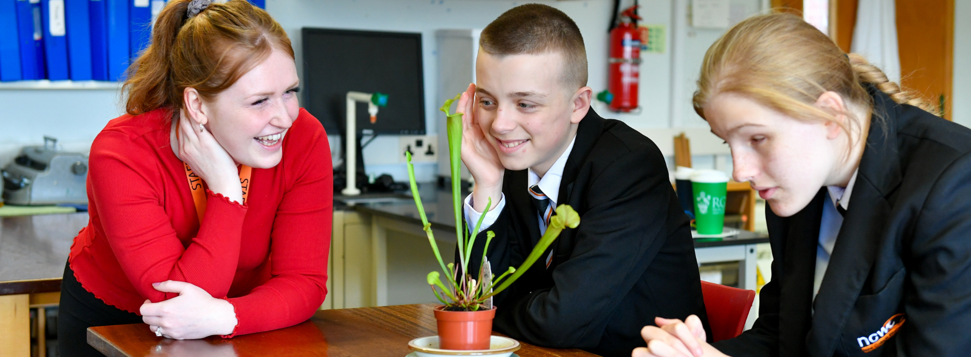 Three students and a member of staff gather around a small potted carnivorous plant during a science lesson. The group leans in closely to observe the plant's distinctive tube-shaped leaves while discussing its features. The classroom contains science equipment, storage shelves and educational resources, creating a hands-on learning environment.