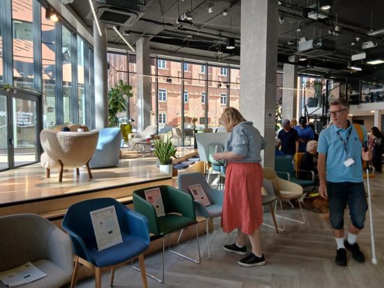 Visitors walk around a modern furniture showroom evaluating seating options during an accessibility consultation event. Various chairs in blue, green, cream and grey are displayed throughout the open-plan space, with information sheets placed on many of the seats. Large floor-to-ceiling windows, indoor plants and breakout seating areas create a bright and spacious environment. A guide dog can be seen resting near a group of attendees in the background.