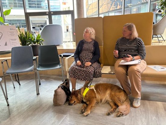 Emma Williams sits in a modern showroom testing furniture as part of an accessibility consultation event. Beside Emma, another attendee makes notes while a guide dog wearing a harness lies comfortably on the floor in front of the seating area. A range of chairs, booths and waiting-area furniture can be seen around the space, which is filled with natural light from large windows.