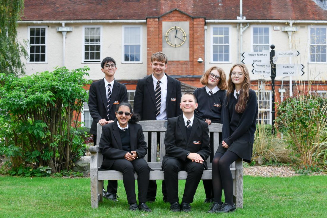 Six students in school uniform are gathered around a wooden bench in the grounds of a school campus. Three students sit on or beside the bench while three stand behind it. The group is positioned on a lawn surrounded by shrubs and ornamental grasses, with a large school building in the background featuring a prominent clock. Several directional signs are visible near the building, pointing to campus facilities and accommodation areas. The image is taken outdoors in daylight and shows the students facing the camera in a group portrait.