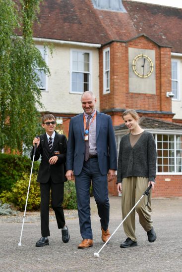 Two NCW students using white canes walk alongside Mr Stephens outside the main NCW building, with its large clock visible above them.