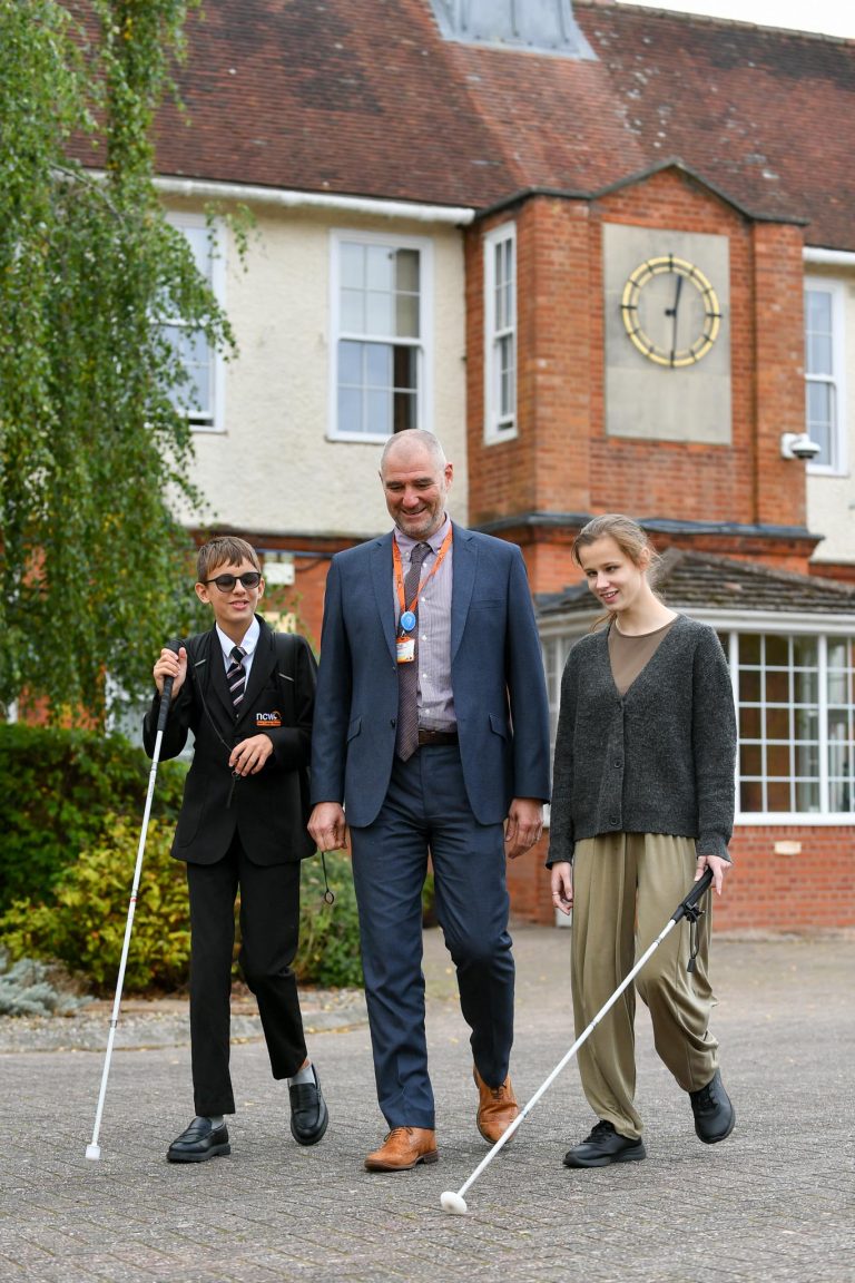 Two NCW students using white canes walk alongside Mr Stephens outside the main NCW building, with its large clock visible above them.