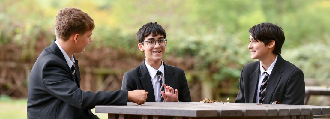 Three students in school uniform sit around a wooden picnic table outdoors, engaged in conversation. Trees frame the top of the image, with a blurred green landscape and distant hills in the background. One student faces the camera while the other two sit opposite each other, creating a relaxed scene in a garden setting.