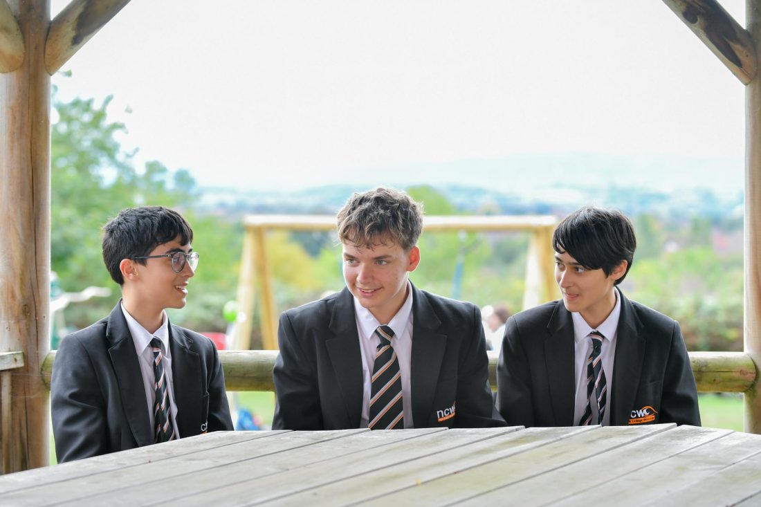Three students in school uniform sit together under a wooden outdoor shelter, gathered around a large picnic table. The students face one another in conversation, with a playground swing frame and a soft-focus green landscape visible in the background. The image is brightly lit by natural daylight, creating a calm outdoor school setting.