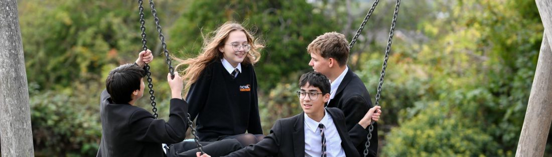 Four NCW students in school uniform gather on a large outdoor basket swing, surrounded by trees and greenery.